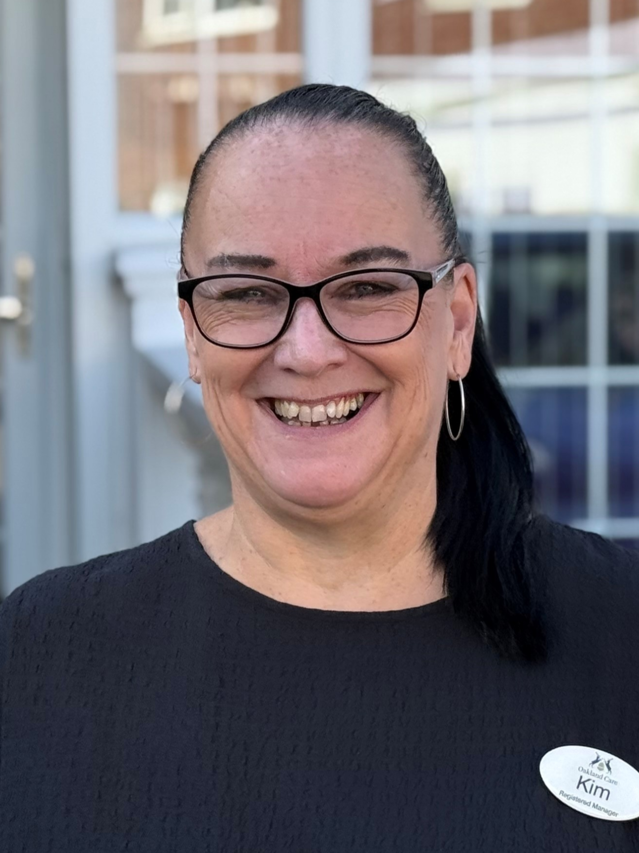 Professional headshot of Kim Maw, a smiling woman with dark hair pulled back, wearing glasses and a black shirt with a name badge.