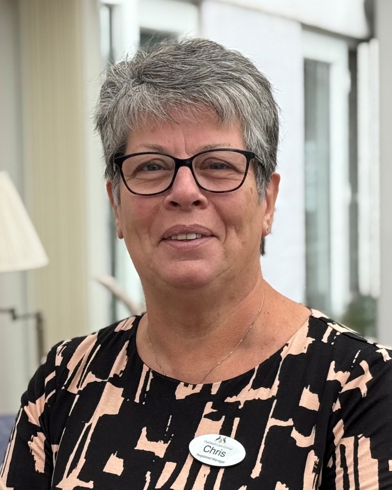 Professional headshot of Chris, a smiling woman with short grey hair and glasses, wearing a black and beige patterned top and a name badge.