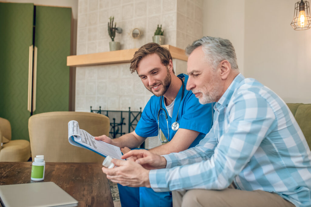 A cheerful physician talks with a serious-looking older man in a medical office. The doctor is explaining something, suggesting a discussion focused on preparing for future care needs or transitioning to a care home.