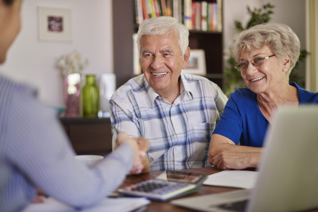 A close-up of two people shaking hands over a desk with documents, a laptop, and a calculator, symbolizing a successful financial agreement or support arrangement, possibly related to elderly care or financial planning.