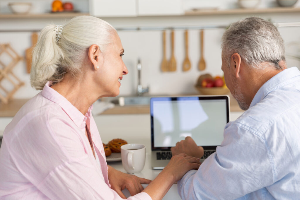 A happy, mature couple sits together in a cozy kitchen, smiling while using a laptop. The scene suggests they are researching or planning for the future, possibly exploring care home options or making financial preparations.
